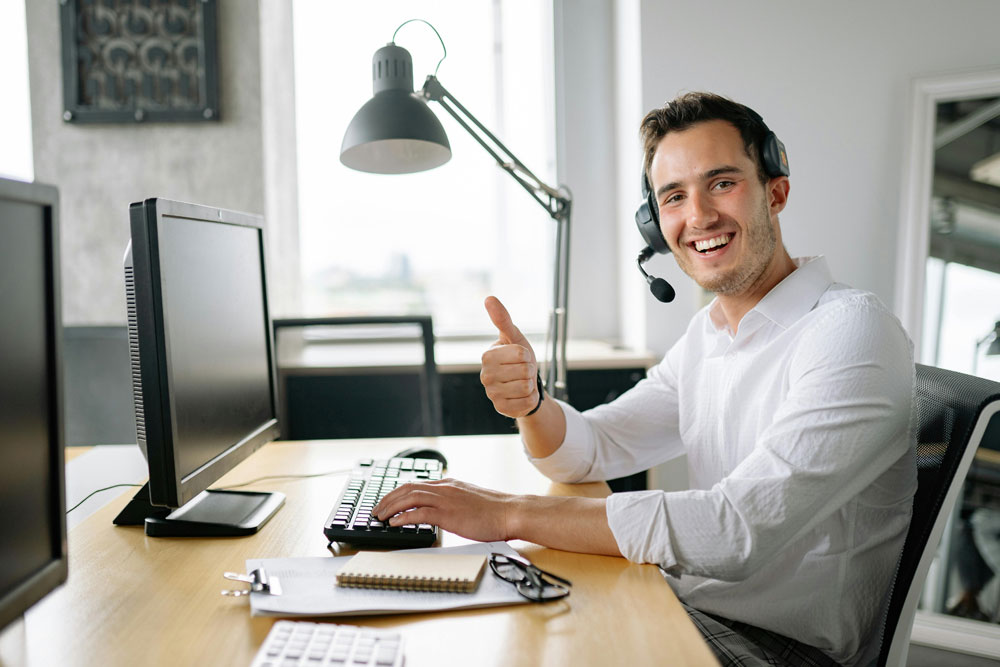 young man smiling into camera with thumb up in office wearing headset